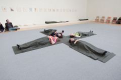 Four people laying down on the floor, each on one leg of a large felt 'X,' with their heads in the center. Each person is covered with a blanket of the same dark gray felt.