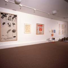 A long white wall with multiple framed works, the largest being a canvas with women's hairstyles painted in various stages of completion with a purple background partially scribbled in, and a painting or drawing of a Campbells soup can, also with scribbled gray and red lines to its left and above.