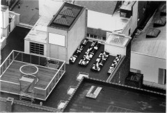 Black and white aerial view of the rooftop of a building with a large rectangular glass-walled sculpture with a cylindrical interior glass sculpture on the left, and four rows of chairs covered in graphic fabric on the right of the images.