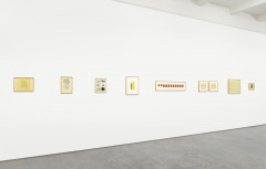 A diagonal view of a white wall with many posters, flyers, and drawings of varying sizes, arranged in a central row. Many are on white paper yellowed with age, and many include black text with illustrations, or blocks of yellow or red.