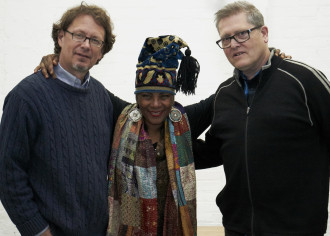 A smiling woman wearing a patterned scarf and tasseled hat poses with her arms around the shoulders of two smiling men wearing glasses in front of a white wall.
