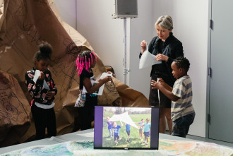 Three children stand in front and next to a Dia educator and hold pieces of printed paper with a large draped sheet of brown drawn on paper in the background and a monitor with an image of playing children sits on a table in the foreground.