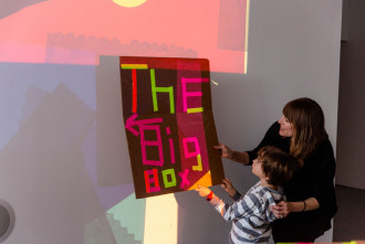 An adult and a child both hold up a cardboard sign reading, “THE BIG BOX” in front of projected light with an arrow pointing out of view.
