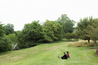 Two people sit on a small grassy hill which overlooks a small grass field that is dotted with trees in various size in the background.