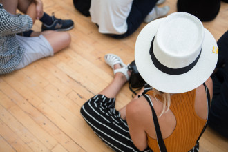 A person in a white hat with and summer clothing sits on the floor in a group of people facing away from the camera.