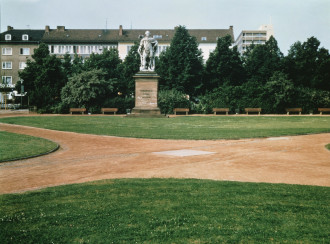 A grassy park featuring four red-dirt paths that intersect at a square silver plate flush to the ground.