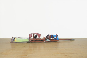 A long, low-lying sculpture made of red, blue, green, and white metal parts rests on a wooden floor.