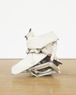 A crumpled-and-folded, white, metallic object with red details rests on a wooden floor in front of a white background.