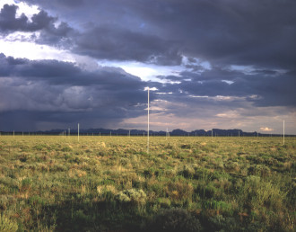 Tall silver poles sticking up out of a grassy landscape, with purple storm clouds in the sky. 