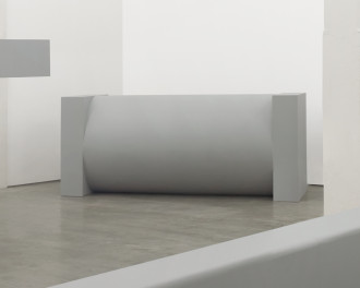 A large, cylindrical, gray sculpture lies lengthwise between two large, rectangular, gray ends.
