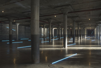 Many thin, long, neon blue lights are evenly scattered across the concrete floor of an industrial basement. Some lights are attached to the background walls.