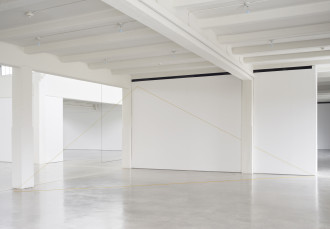 A yellow thread is suspended from the ceiling and runs along the floor, creating a triangle in a white industrial space. A second work in dark thread is partially visible farther into the background.