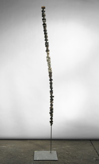 Balanced on a bronze pole and attached to a flat base is a thin vertical sculpture made of several painted corks stacked on top of one another. 
