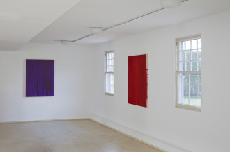 Two paintings, a purple rectangular canvas and a red rectangular canvas and a on adjacent white walls between two windows.