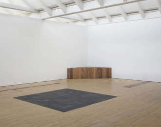Four sculptures in a the corner large gallery with wood floors and white walls, including copper squares in a row coming from the wall, a large dark square in the middle of the space, and a triangular stack of wood in the corner.