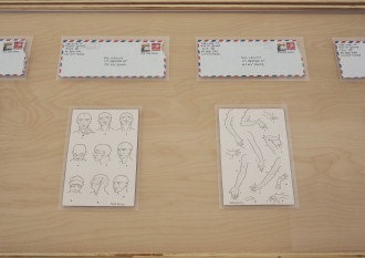 Several envelopes addressed to Sol Lewitt are placed on a wooden surface above two drawings that respectively feature many angles of a person's head and arm.
