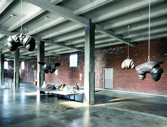 Four phallically shaped, metallic objects are suspended by wires from a cement ceiling in the foreground of a large, warehouse-like room with brick walls, cement columns, and cement floor. A large, low table with many organically shaped objects is centrally placed in the room.