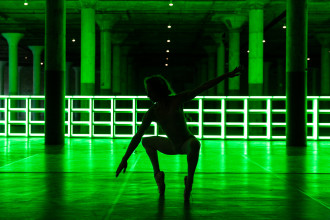 A dancer crouches en pointe facing forward with their face in left profile, right arm extended diagonally upwards and left arm extended diagonally downwards. They are backlit with a neon green, gridded light installation that runs the length of the floor in the background.