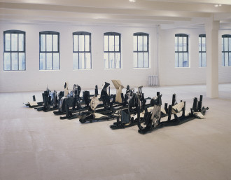 Seven rows of black steel frames sit on the ground in an empty white space with windows. Black and white crushed automobile steel forms of different sizes and shapes extend upwards from the frames.