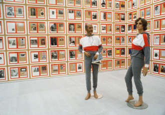 Two mannequins in identical white, red, and grey tracksuits, posed in front of a floor-to-ceiling locked grid of paper works.