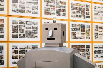 The top half of a square-shaped, grey robot angles forward. Its eyes and mouth are horizontal rectanglar holes and its nose is a protruding triangular prism. The visible leftside of the head has a circle hole in the place of an ear. The top of head has a protruding lit up lightbulb. The background wall is completely covered in framed images.