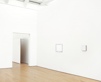 Two square white paintings hang on either side of a corner; the left one is very large and the right one is very small. A large, rectangular white painting affixed to a wall by long metal rods and resting on the wood floor is partially visible through a doorway to the left of the paintings.