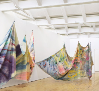 Two massive swathes of multicolored canvas are tied at various points and strung up from the ceiling of an airy, bright, white room. The canvases are painted and splattered with an array of vibrant, pastel, and metallic colors. The artwork floats above the floor. The swathe of canvas in the foreground closely hugs the wall and the canvas in the background arcs outwards with a corner pinned to the wall.