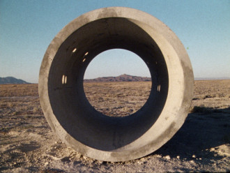 A large, concrete tube oculus looks out onto a desert basin, a faraway mountain range, and a clear blue sky. The tube has various sized circles drilled through the concrete in a constellation pattern and outside light casts into the space.