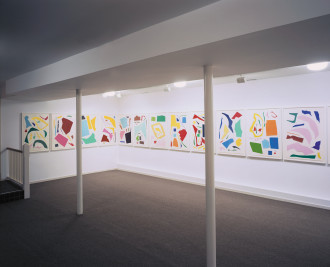 Twelve framed colorful works made up of abstract cutouts hang close together on a white wall in a low-ceilinged room.