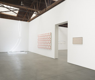 A thin, neon white string of fluorescent light hangs from the exposed ceiling in two strands from a single point. Two paintings of identical geometric subject matter but different scale hang on the wall with a doorway between them.
