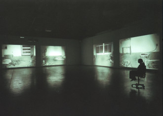 A lone figure sits on a chair in a large, dark room and looks out at four night vision projections of different rooms.