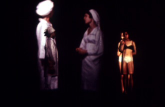 An out of focus image of three people. Two people against a wall have images of other people projected onto them. The projection on the left is dressed in all white and the projection on the right is dressed in a black bra and white underwear. A third person dressed in white stands away from the wall in the middle of the two other people.