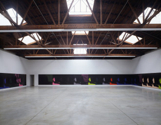 Identical multicolored paintings line the entirety of a gallery room in large open space with three walls depicted.