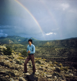 A man stands on a rocky terrain with a low mountain range behind him and a double rainbow in the sky.