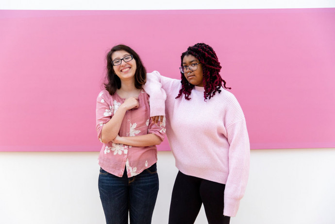 Two adults, both wearing pink tops, stand casually posed in front of a purely pink color field that occupies the top two thirds of the image.