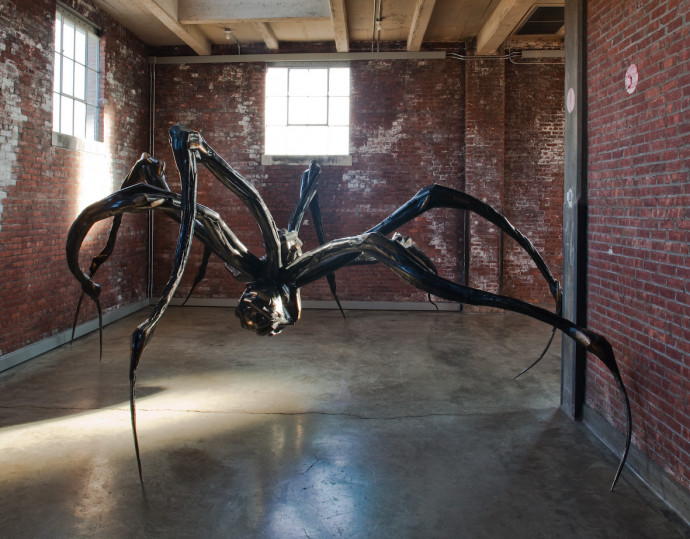 Large bronze spider sits on cement floor in front of two brick walls with windows.