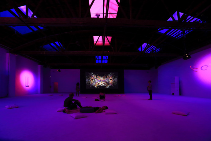 An industrial space is bathed in blue, pink, and purple light with skylights overhead. Five people stand, sit, or lay on small rectangular pillows on the floor. There are three minimal sculptures on the walls and a colorful abstract projection on the back wall.  