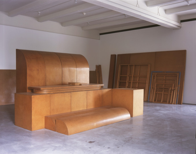 Large, smooth, curved wooden shapes and cubes abutt and pile atop each other in the center of a room, with wooden panels and empty frames leaning on the walls, all stained a reddish brown.