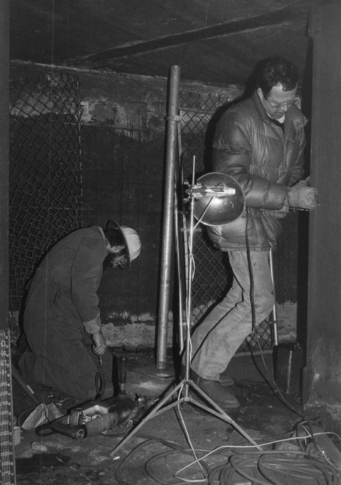 Two men are working with tools in an enclosed dark space in this black-and-white image.