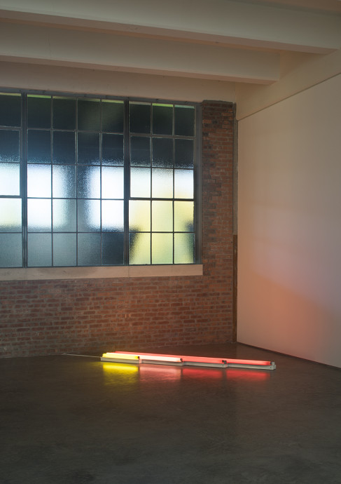 Four neon tubes of increasing length lie beside one another on the floor in the corner of an industrial gallery space. The shortest tube is gold, the next is pink, and the two longest are both red.
