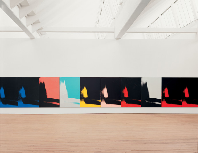 Ten canvases hang edge to edge and low to the floor. Each canvas features an identical abstract mark painted in black, gray, red, green, yellow, peach, or blue. 